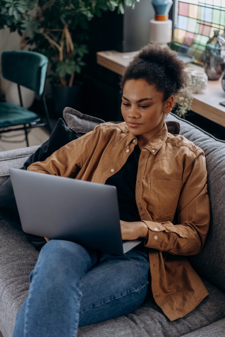 Woman in Brown Jacket and Blue Denim Jeans Sitting on Gray Couch Using Macbook